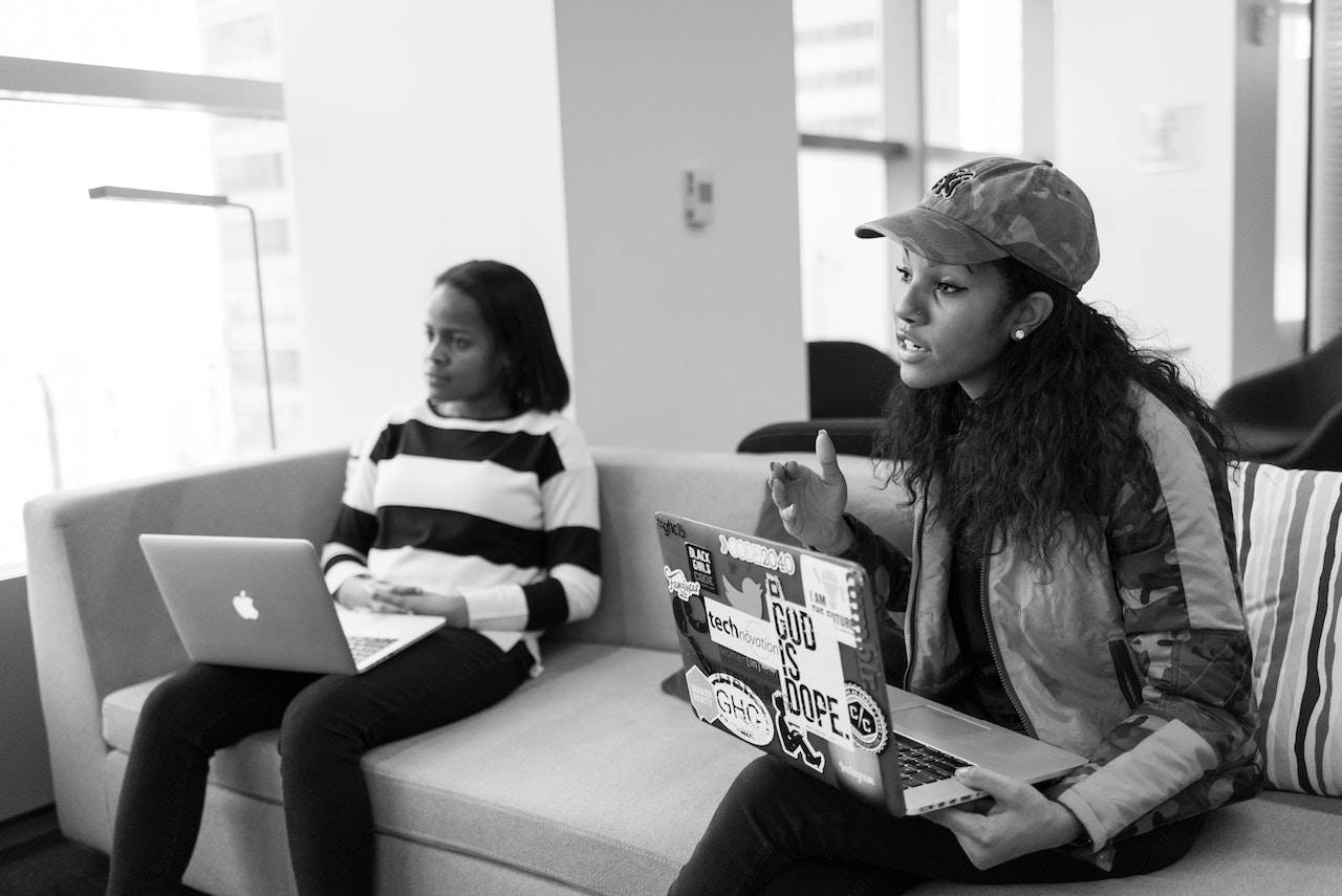 Two black women sit on a sofa holding laptops